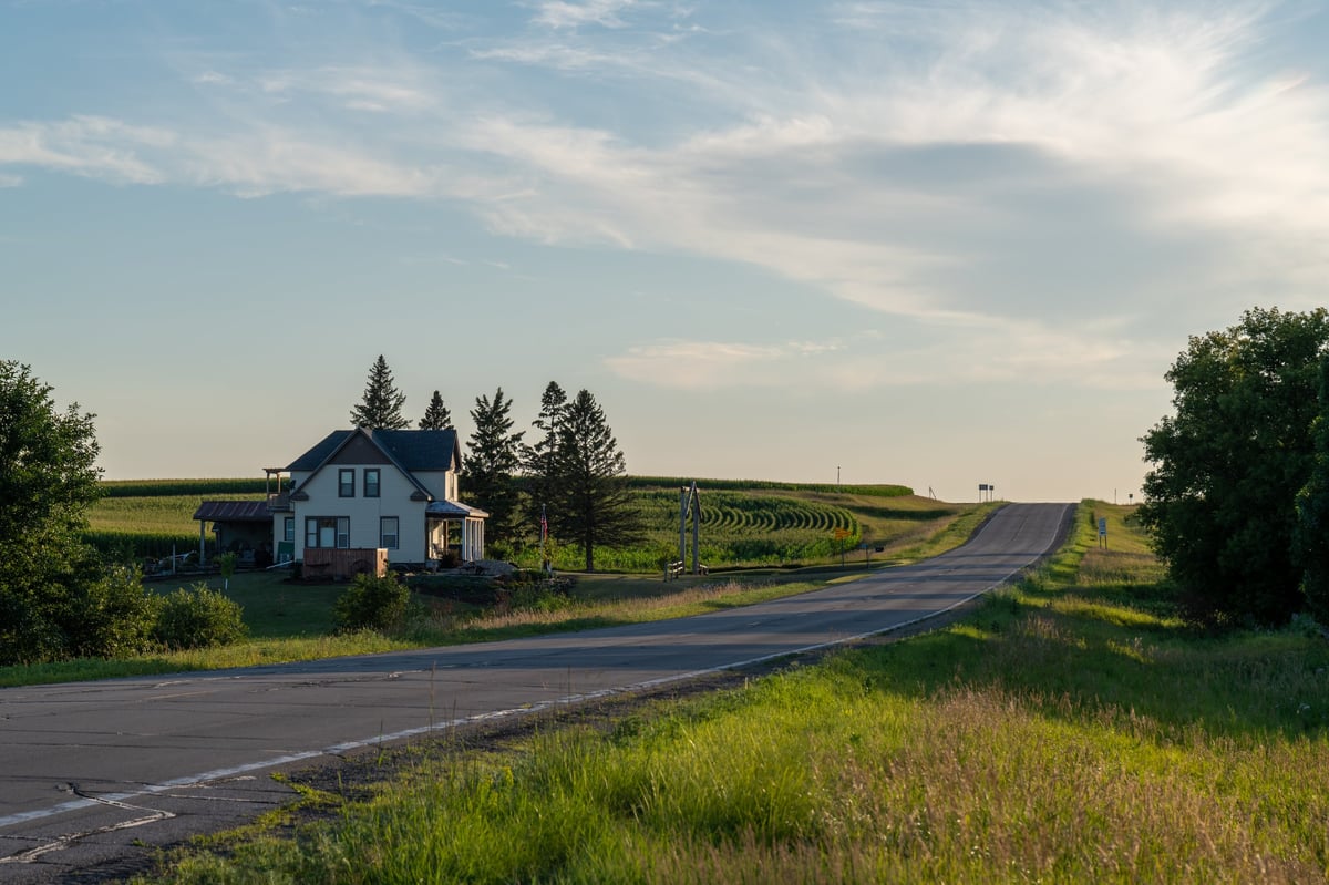 Looking down a country road lined with fields and a white farmhouse on a calm evening in rural Minnesota, USA.