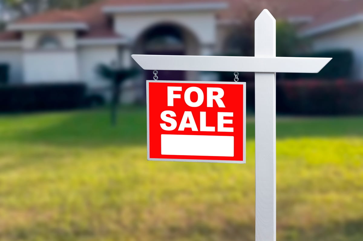 A sign stands in the grass with the words for sale on it. The sign marks a house that is available for purchase.